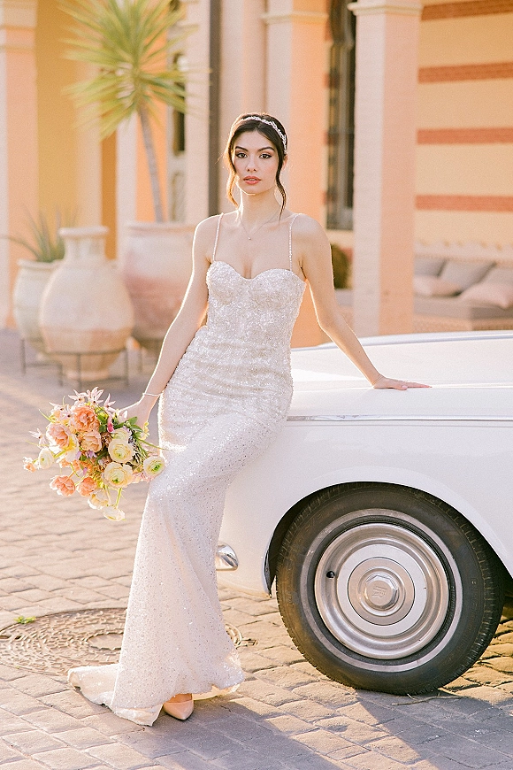 Bridal portrait of a bride in a sparkly wedding dress holding a peach and cream bouquet beside a vintage car in a palm courtyard