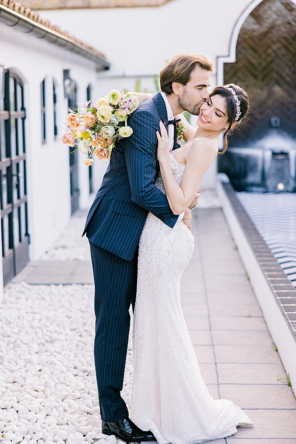 Couple portrait of groom kissing bride’s forehead as she holds a pastel bouquet, standing by a white arched villa doorway outdoors
