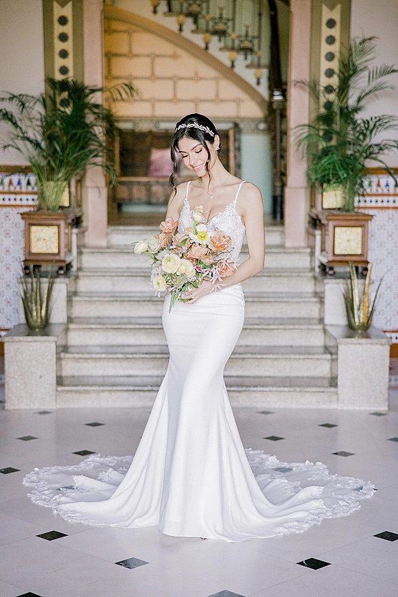 Bridal portrait of a bride in a wedding dress with train, holding a pastel bouquet on a grand staircase beneath a chandelier
