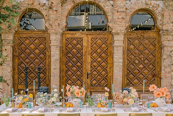 Reception tablescape with long banquet table decor, pastel floral centerpieces, taper candles, and citrus accents under string lights by brick arches
