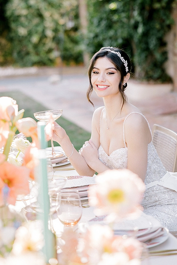 Bridal portrait of a smiling bride at reception table in a sparkly gown, holding a champagne coupe in a sunlit garden setting