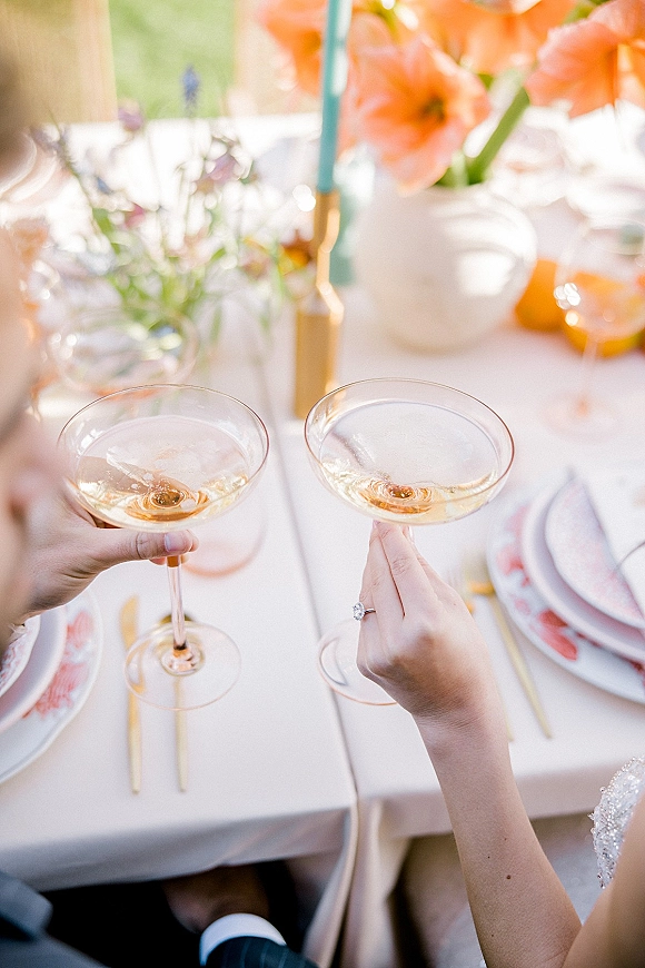 Wedding toast as hands clink coupe champagne glasses, engagement ring visible beside a taper candle and floral centerpiece on a lawn table