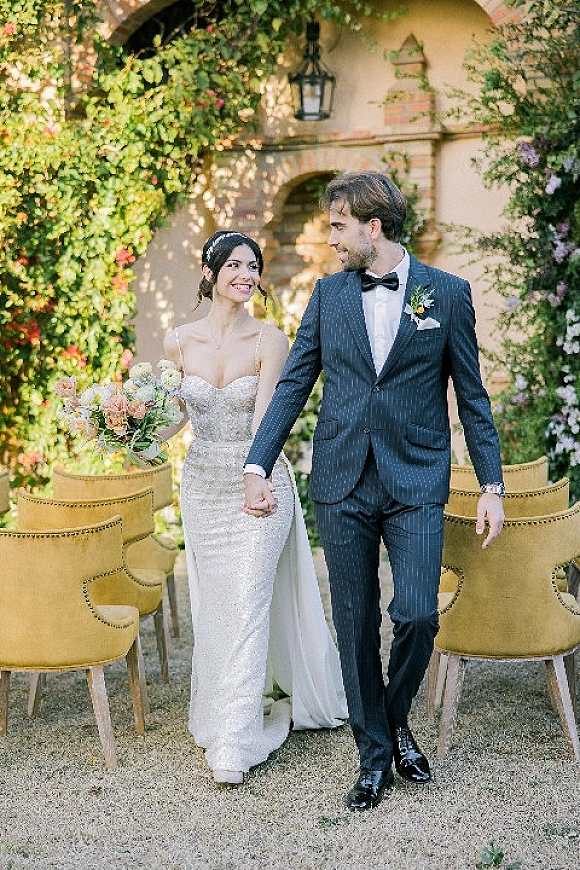 Couple portrait of bride and groom holding hands, bride gazing at him, with bouquet accent by vine-covered stone archway walkway