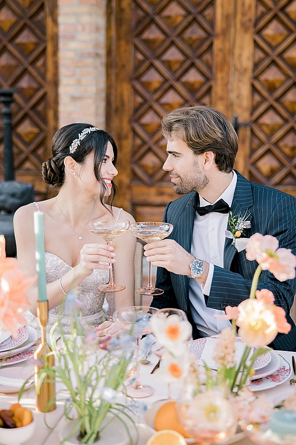 Wedding couple toast with champagne coupe toast, clinking glasses at a candlelit sweetheart table before wooden doors and brick wall