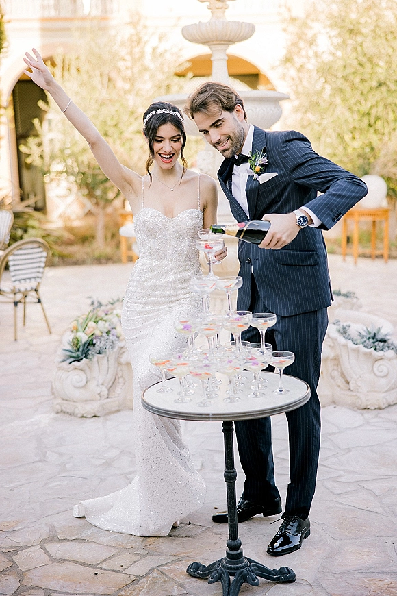 Champagne tower moment as bride and groom in courtyard pour bubbly into coupe glasses on a pedestal table, fountain behind