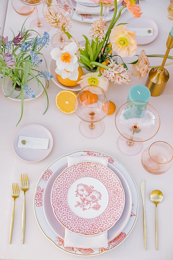 Reception tablescape featuring a wedding place setting with patterned plates, gold flatware, pink stemware, citrus, florals, and a taper candle on white cloth