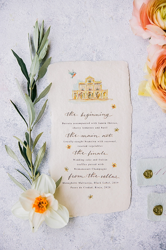 Wedding menu card with wedding dinner menu in calligraphy on deckled paper, styled with ribbon and wax seal on a light gray stone surface