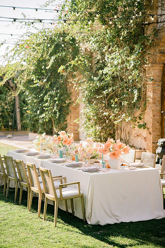 Reception tablescape with peach floral centerpieces and taper candles on white linens, set under string lights beside ivy walls and brick arches