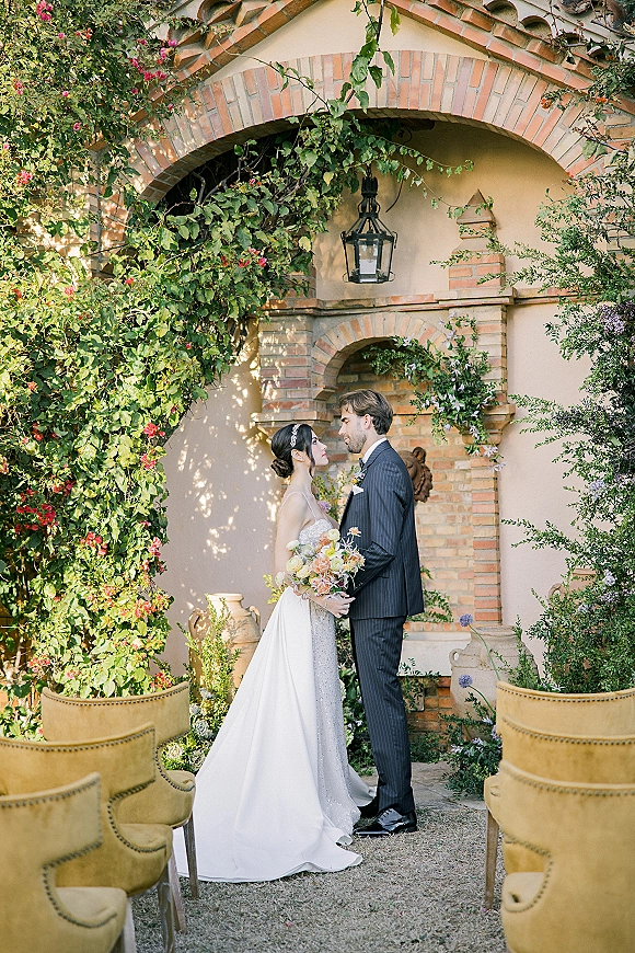 Couple portrait of bride and groom standing face to face, her bouquet and long veil in a courtyard with brick arches and vines