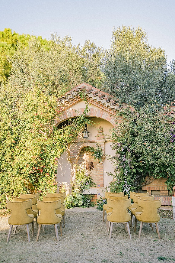 Ceremony setup for an outdoor wedding ceremony with mustard velvet chairs, greenery garland, and lanterns framing a brick arch and fountain