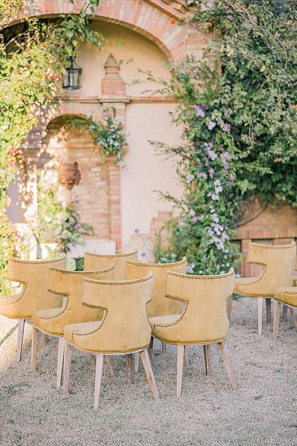 Ceremony seating with outdoor ceremony chairs in mustard velvet, nailhead trim details, set on a lawn before a brick arch wall with vines