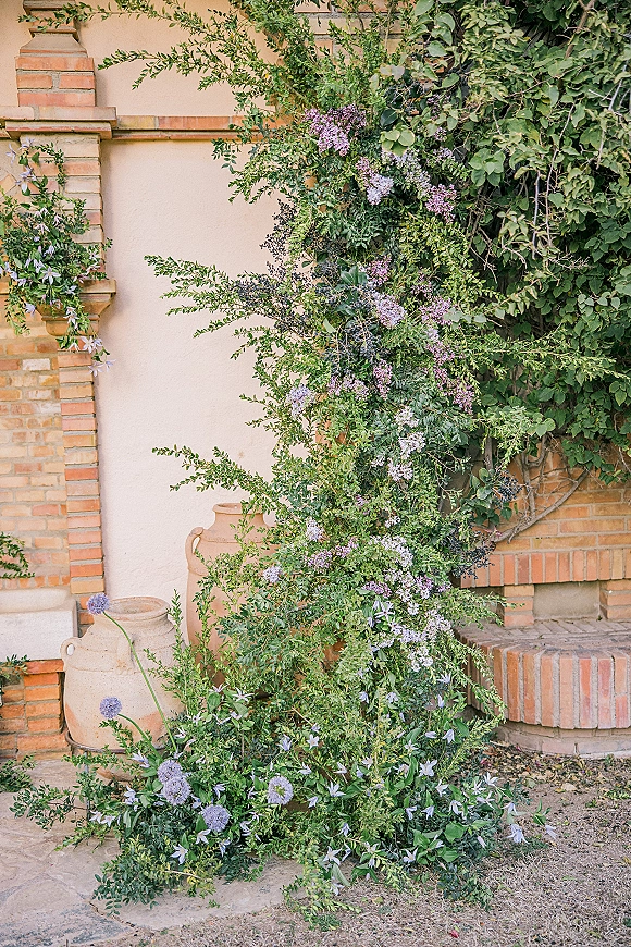 Wedding floral installation with lavender blooms and airy greenery draping ceramic urns against a brick wall in an outdoor courtyard setting