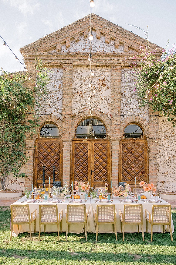 Reception tablescape with an ivory-draped long dining table, cane-back chairs, floral centerpieces, taper candles, and string lights in a vine-covered courtyard