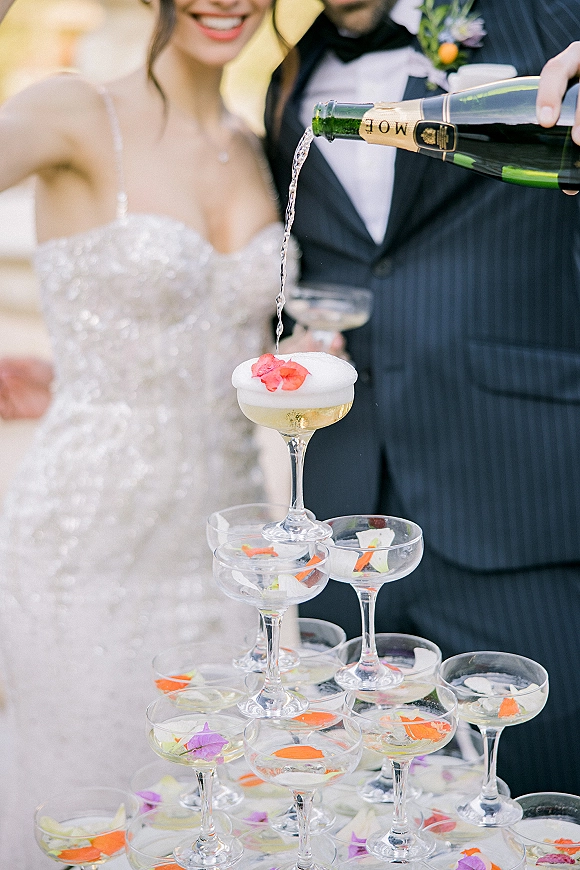 Champagne tower setup with coupe glasses and a champagne bottle, sprinkled with edible flower petals as the couple blurs in greenery behind