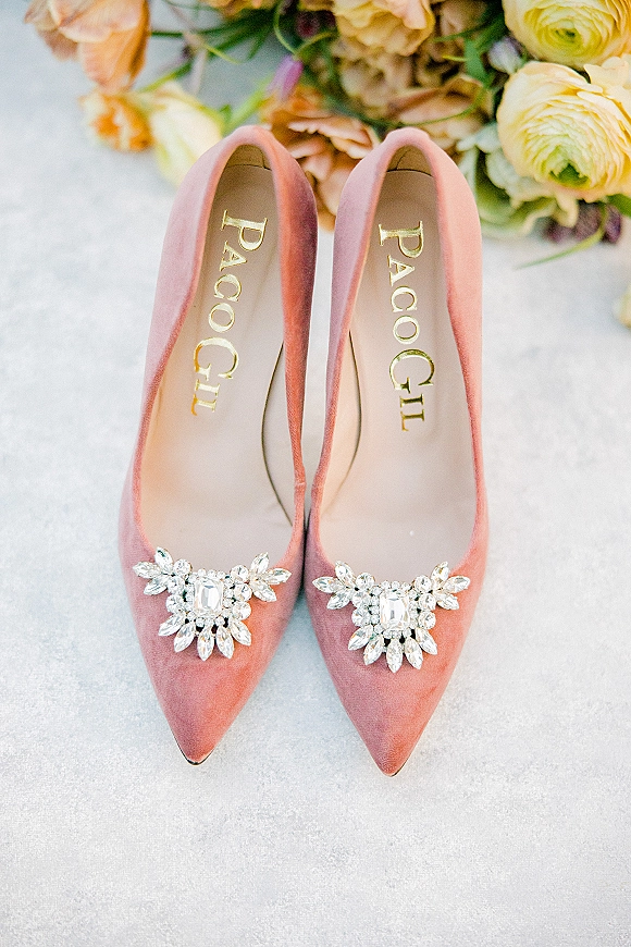 Wedding shoes in blush velvet heels with crystal shoe clips, arranged on a light stone surface beside flowers