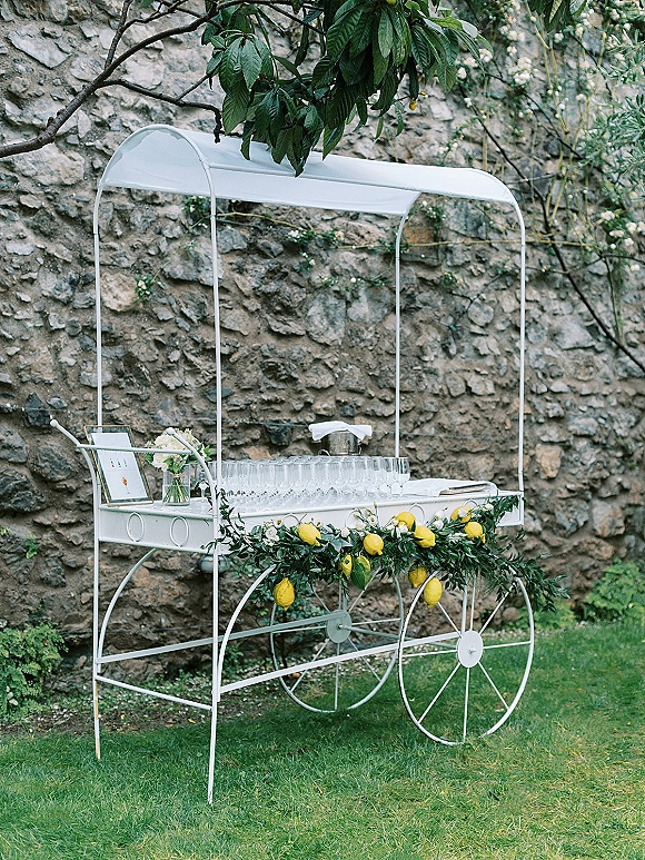 Wedding drink cart with champagne cart wedding setup, lemons, flutes, and greenery garland under a canopy beside a stone garden wall
