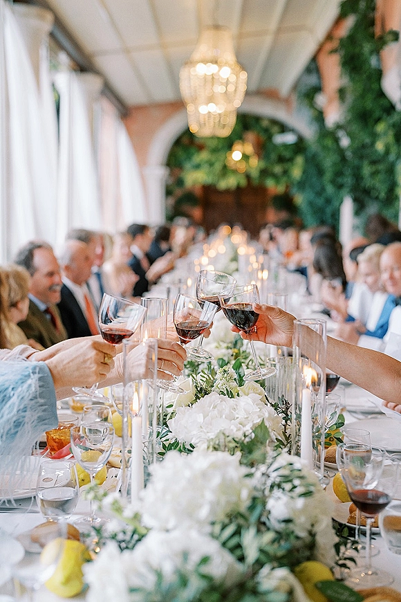 Wedding reception toast with guests cheering, clinking red wine glasses over a long candlelit banquet table beneath a chandelier