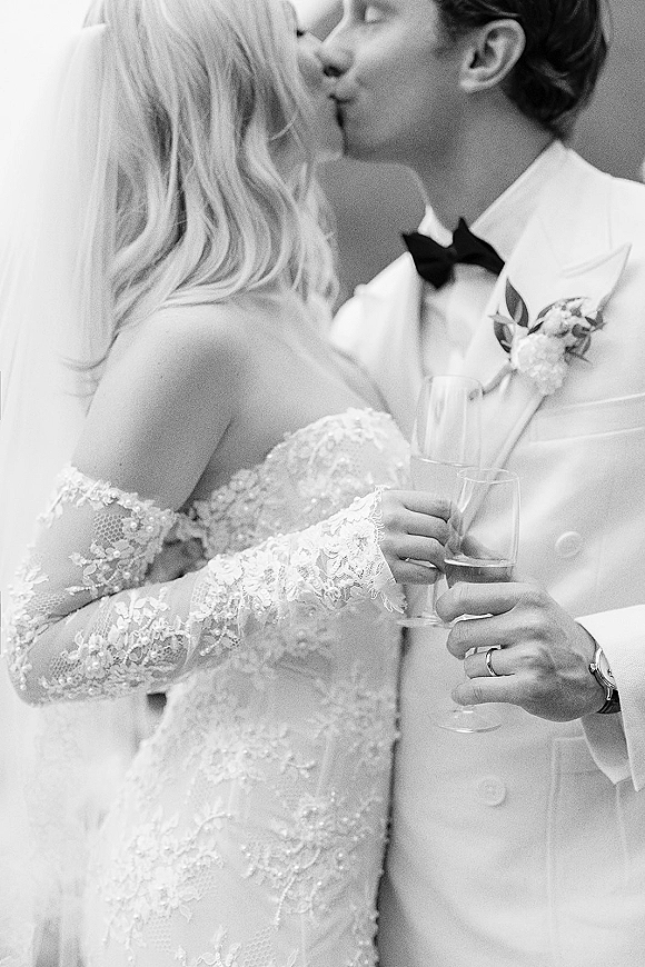 Wedding kiss portrait of bride and groom kissing, each holding champagne flutes; her lace sleeves and veil meet his bow tie by an indoor wall