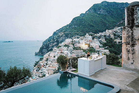 Wedding reception bar with a white counter and chandelier, set on a terrace beside an infinity pool with ocean and mountain views