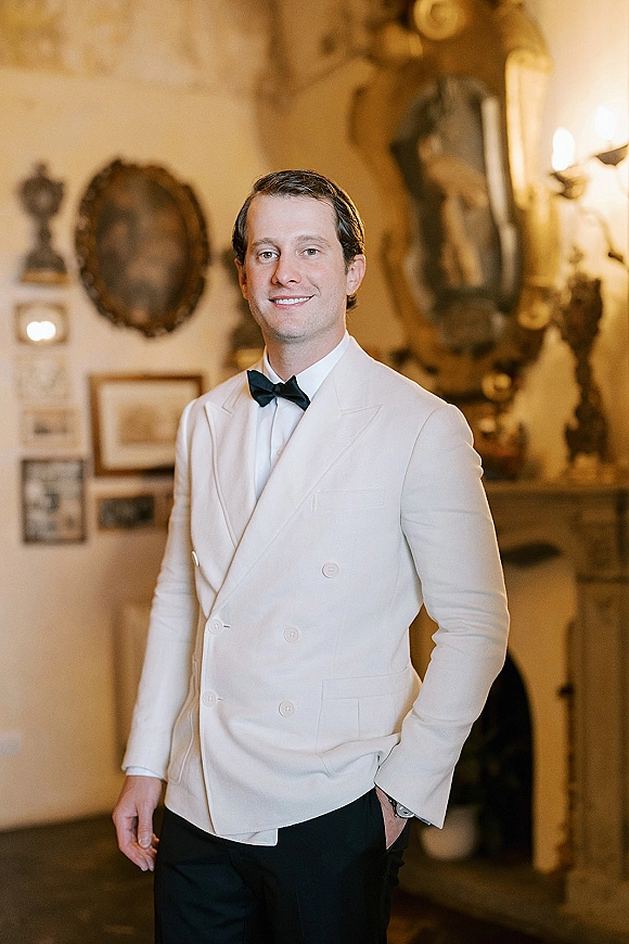 Groom portrait in a white tuxedo jacket with black bow tie, hands in pockets, standing in an indoor room with mirrors and sconces