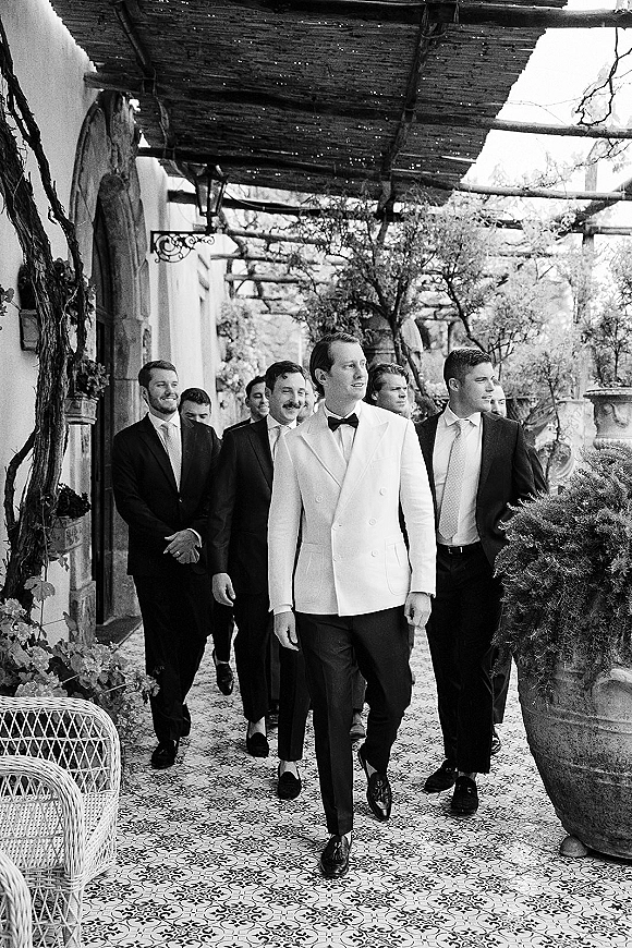 Groomsmen photo of men walking together in coordinated suits and ties, including one in a white dinner jacket on a covered patio with pergola vines