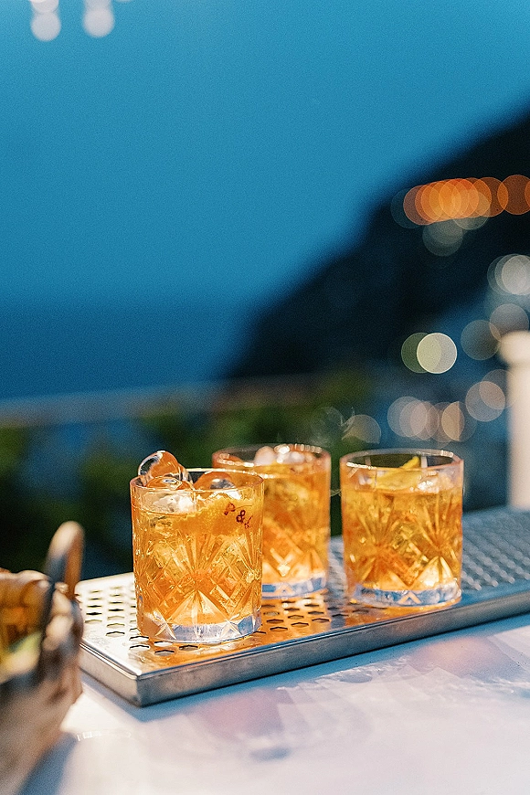 Wedding signature drinks in rocks glasses with amber cocktails, ice cubes and citrus peel garnish on a metal tray under bokeh string lights