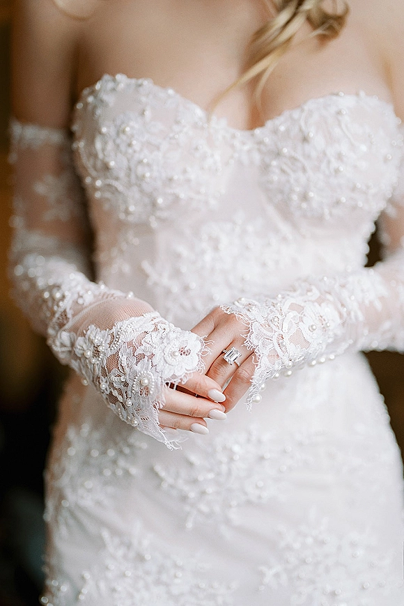 Wedding dress close-up showing a strapless lace wedding dress bodice with pearl beading, lace gloves, and an emerald-cut ring indoors