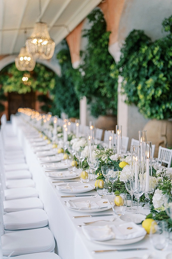Reception tablescape with long head table decor featuring white florals, greenery garland, lemons, taper candles, and chandeliers under a tent canopy