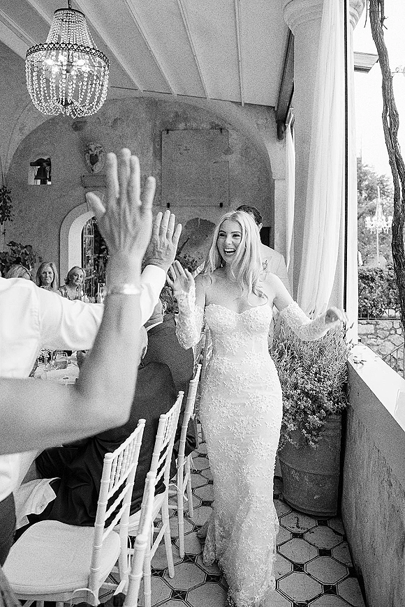 Bride entrance as she laughs and high fives guests in a lace off-the-shoulder gown and veil under a chandelier on a covered patio