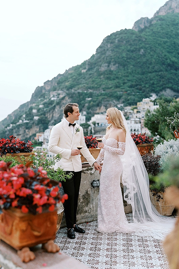 Couple portrait of newlyweds toasting with wine glasses, bride in long veil and groom in white jacket on a tiled terrace by mountains