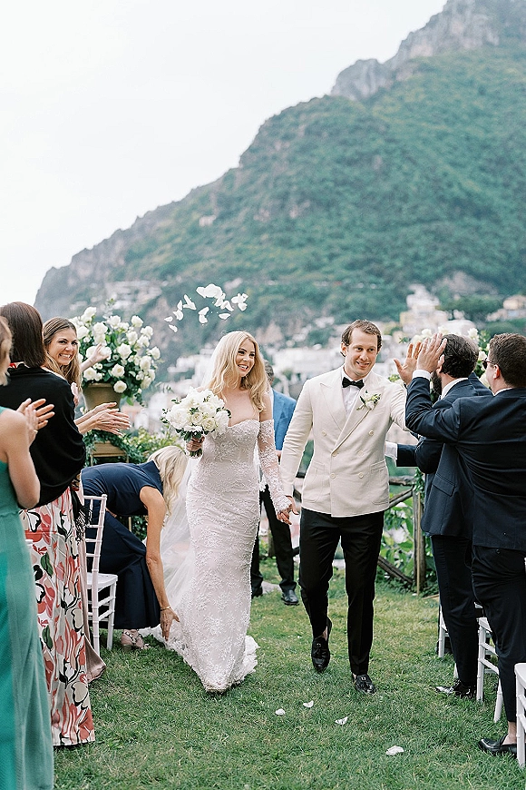 Wedding recessional as bride and groom walk the aisle hand in hand, guests tossing petals, mountains behind white chairs and greenery garland