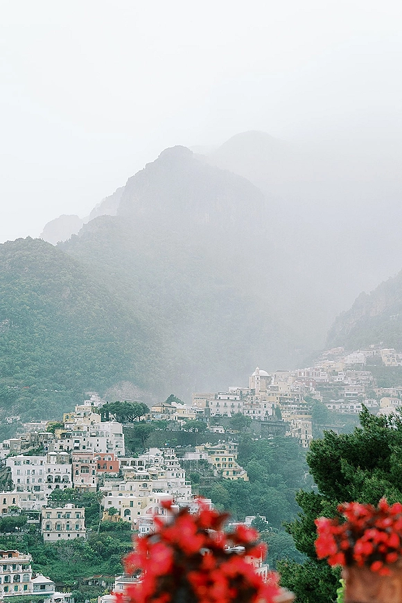 Wedding scenery with red flowers in the foreground overlooking a hillside town, mountain cliffs, greenery, and a misty sky