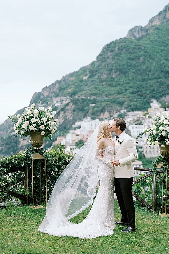 Wedding kiss portrait of bride and groom kissing, her veil blowing as they embrace on a lawn terrace with mountain hillside views