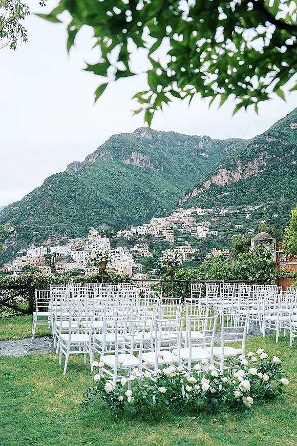 Outdoor ceremony setup with white chiavari chairs lined along an aisle, accented by white rose urn florals on a stone terrace with mountain views