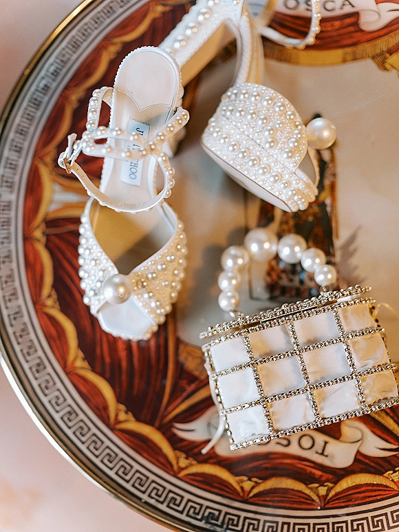 Bridal accessories flatlay with pearl wedding shoes, a beaded hat, pearl necklace, and rhinestone clutch arranged on a tabletop surface