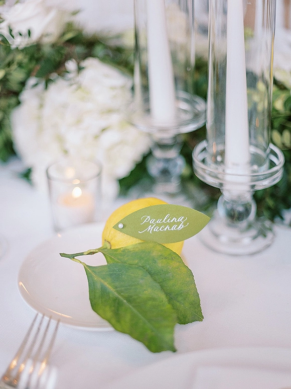 Wedding place card with lemon place card and leaf calligraphy tag beside white taper candles, white florals, and greenery on a reception table setting