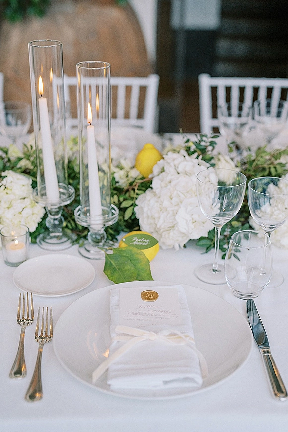 Reception tablescape with white wedding table decor, hydrangeas and greenery garland, tall cylinder candles, lemons, and wax-seal place cards in window light