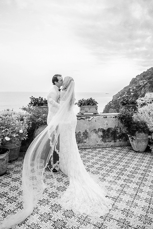 Wedding kiss portrait of bride and groom kissing as her long cathedral veil blows on a terrace with ocean view and cliffs behind