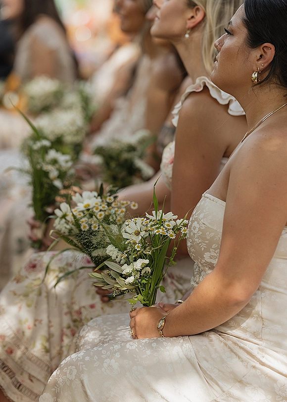 Bridesmaids portrait with wildflower bouquets, seated in warm sunlight by ceremony chairs, wearing floral dresses and gold hoop earrings