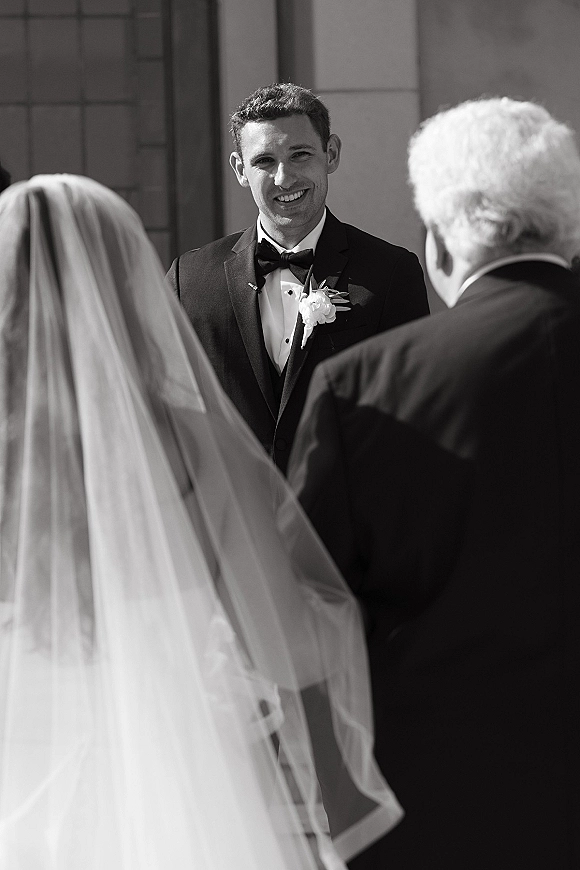 Wedding vows as groom smiles at bride through her bridal veil, wearing a tuxedo with bow tie and boutonniere outside a building exterior