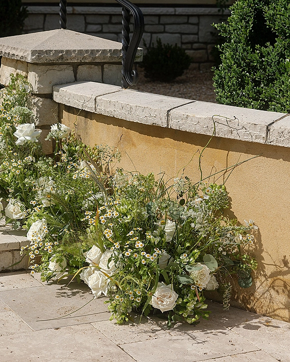 Wedding floral arrangement with white roses, chamomile daisies, and eucalyptus on stone steps beside a stucco wall and wrought iron railing