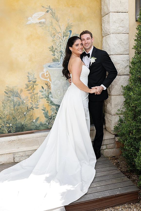 Couple portrait of bride and groom holding hands, her strapless wedding dress with long train beside a painted mural wall and stone column