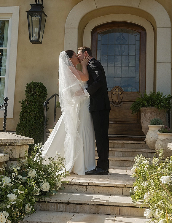 Wedding kiss portrait of bride and groom kissing on stone steps, her veil blowing, by an arched doorway with white rose florals