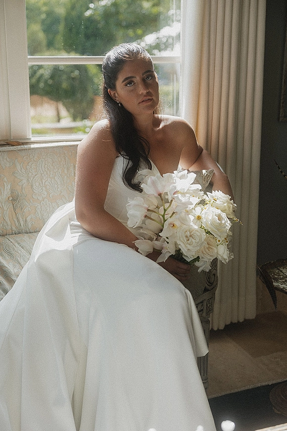 Bridal portrait of a bride in a strapless wedding dress holding a white rose bouquet by a window with curtains and soft indoor light