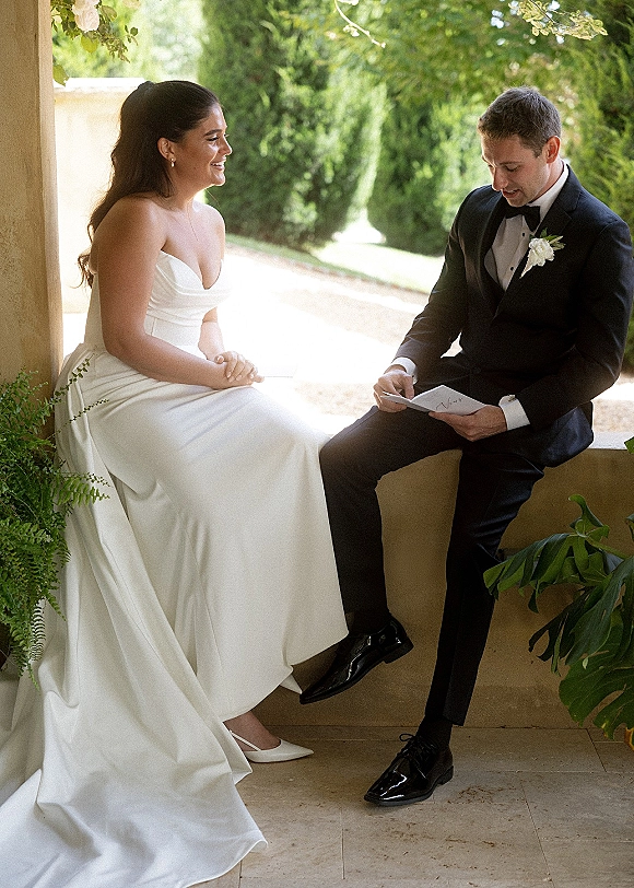 Wedding couple portrait of bride in a strapless dress smiling as the groom reads a note card on a sunlit stone bench patio in a garden