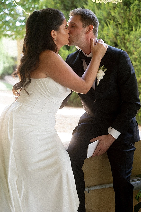 Wedding kiss portrait of bride and groom kissing, her hand on his face, in tuxedo and strapless dress on a tree-lined walkway