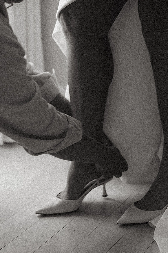 Bride putting on shoes during wedding shoes getting ready, fastening white slingback heels beside her robe in soft window light on wood floor
