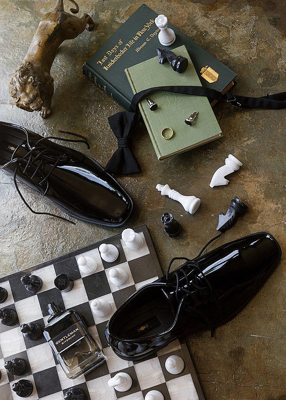 Groom accessories flat lay with black patent dress shoes, bow tie, cufflinks and wedding band beside a chessboard on stone floor