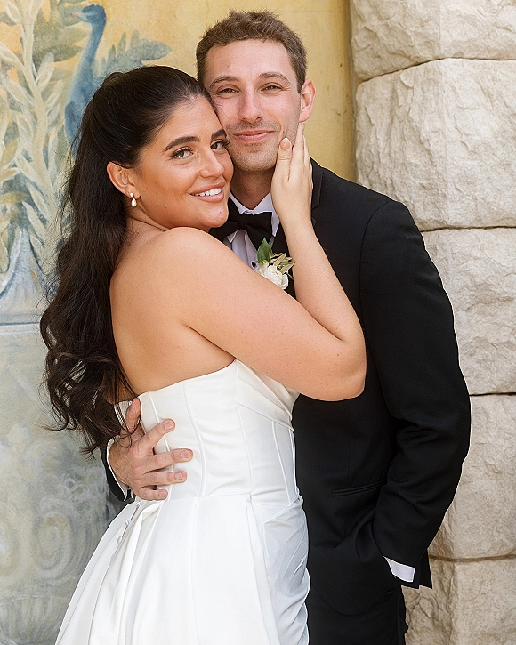 Couple portrait of bride and groom embrace close up, her hand on his face, beside a painted mural and stone wall backdrop