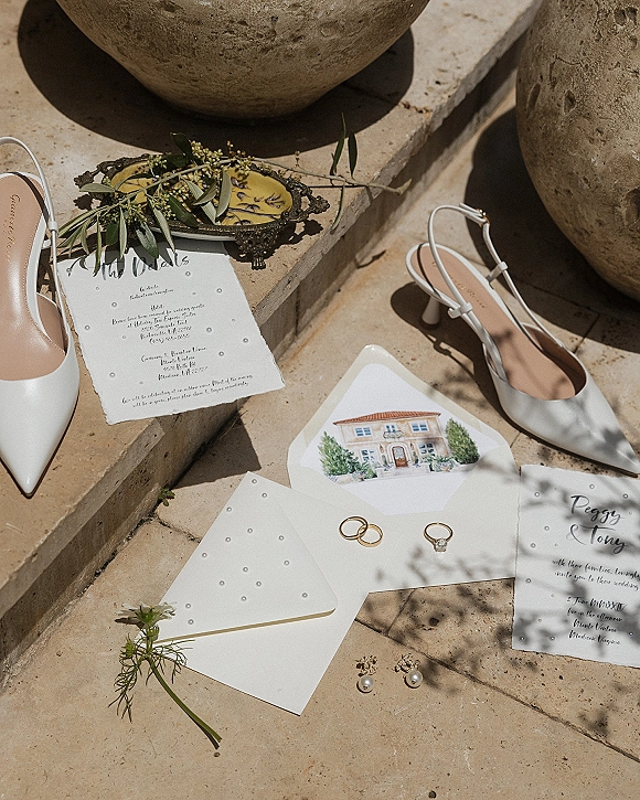 Wedding invitation suite with calligraphy wedding invitation, rings and pearl earrings on a vintage tray beside white slingback heels on sunlit stone steps
